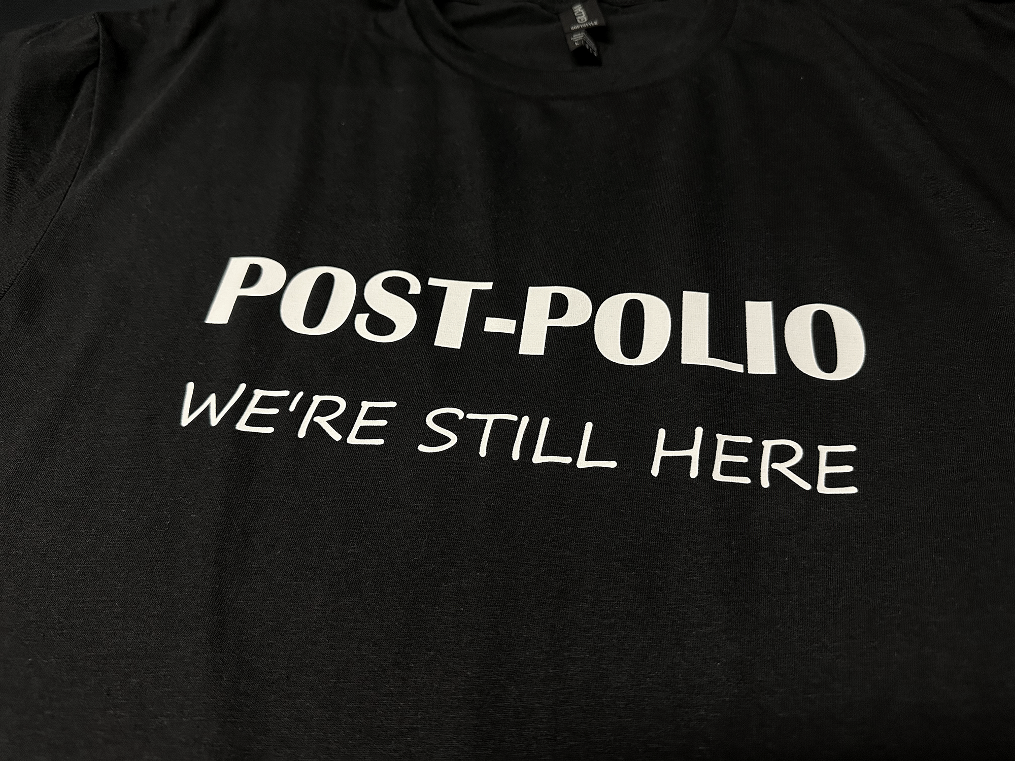 World Polio Day T-shirt in black with white text "POST-POLIO WE'RE STILL HERE" on soft, classic fit fabric.