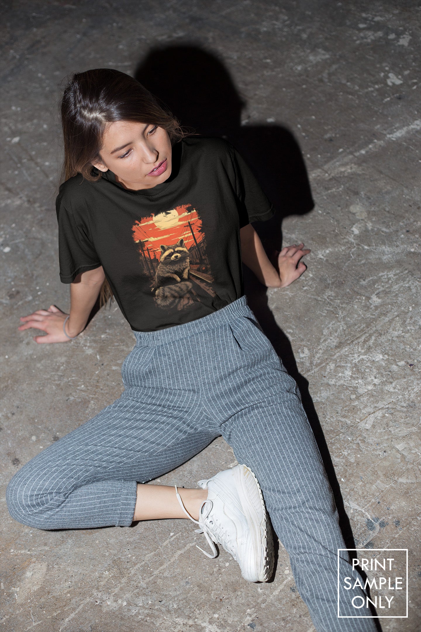Men / Unisex Softstyle 100% Cotton Tee in black with raccoon graphic worn by seated woman on concrete floor.