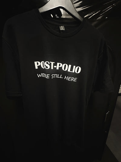 World Polio Day T-shirt in black with white "Post-Polio We're Still Here" text, celebrating polio survivors and community.