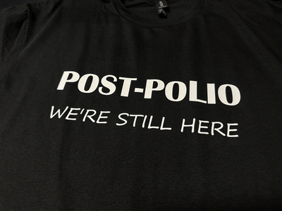 World Polio Day T-shirt in black with white text "POST-POLIO WE'RE STILL HERE" on soft, classic fit fabric.